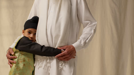 Studio-Shot-Of-Young-Sikh-Boy-With-Top-Knot-Turban-Hugging-Grandfather-Against-Plain-Background-1